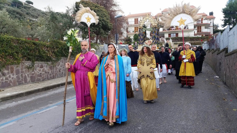 Processione colorata delle Pacchianelle a Vico Equense, con partecipanti in costumi tradizionali del presepe napoletano del XVIII secolo, bambini vestiti da Pacchianelle con cesti di frutta e prodotti tipici della Campania, donne in abiti contadini tradizionali e musicisti che suonano zampogne e ciaramelle, sullo sfondo della Costiera Amalfitana.